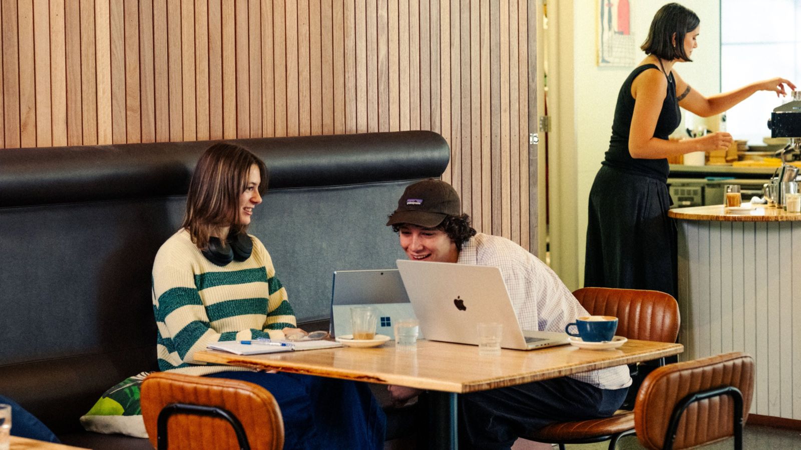 Two students sitting at a cafe table with their laptops, one leaning over to look at something on the other's laptop.