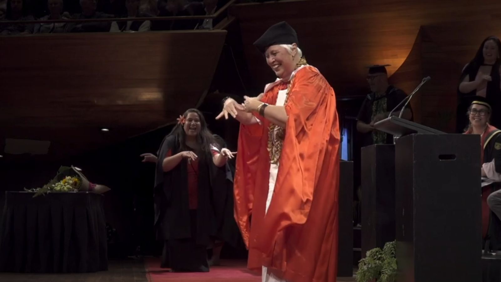 A recipient of an Honorary Doctorate in red regalia dances joyfully on stage during a graduation ceremony, with a graduate smiling and joining in behind her.
