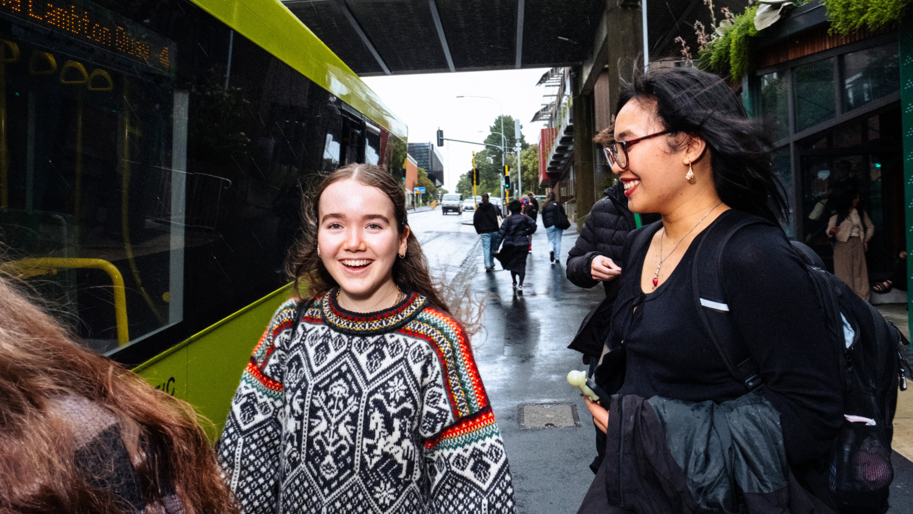 Street scene with pedestrians and a green bus; two people smiling in the foreground—one in a patterned sweater, the other in black with a backpack.