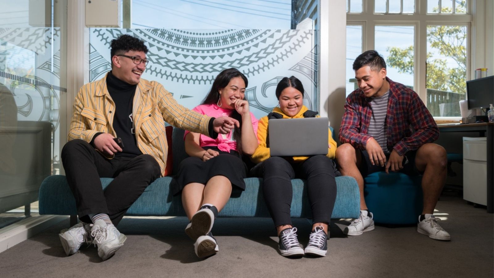Two male and two female students sit on a couch in a Pasifika room. One student in the middle has a laptop on their lap. All the students are looking at the laptop screen and smiling and laughing. One student on the end of the couch is pointing at the screen in mid-laughter.