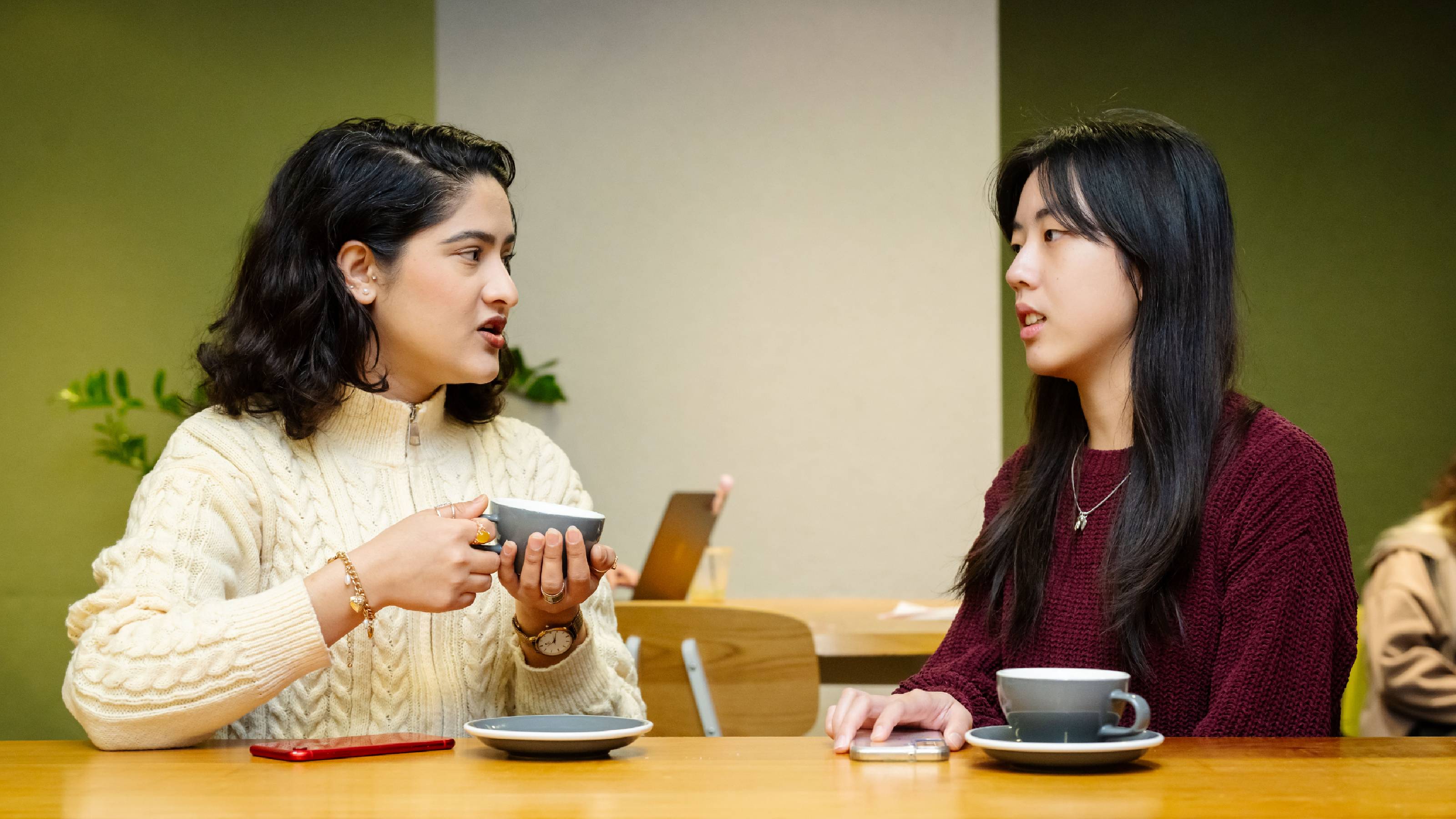 Two students maintaining eye contact as they talk at a table in The Lab over cups of tea