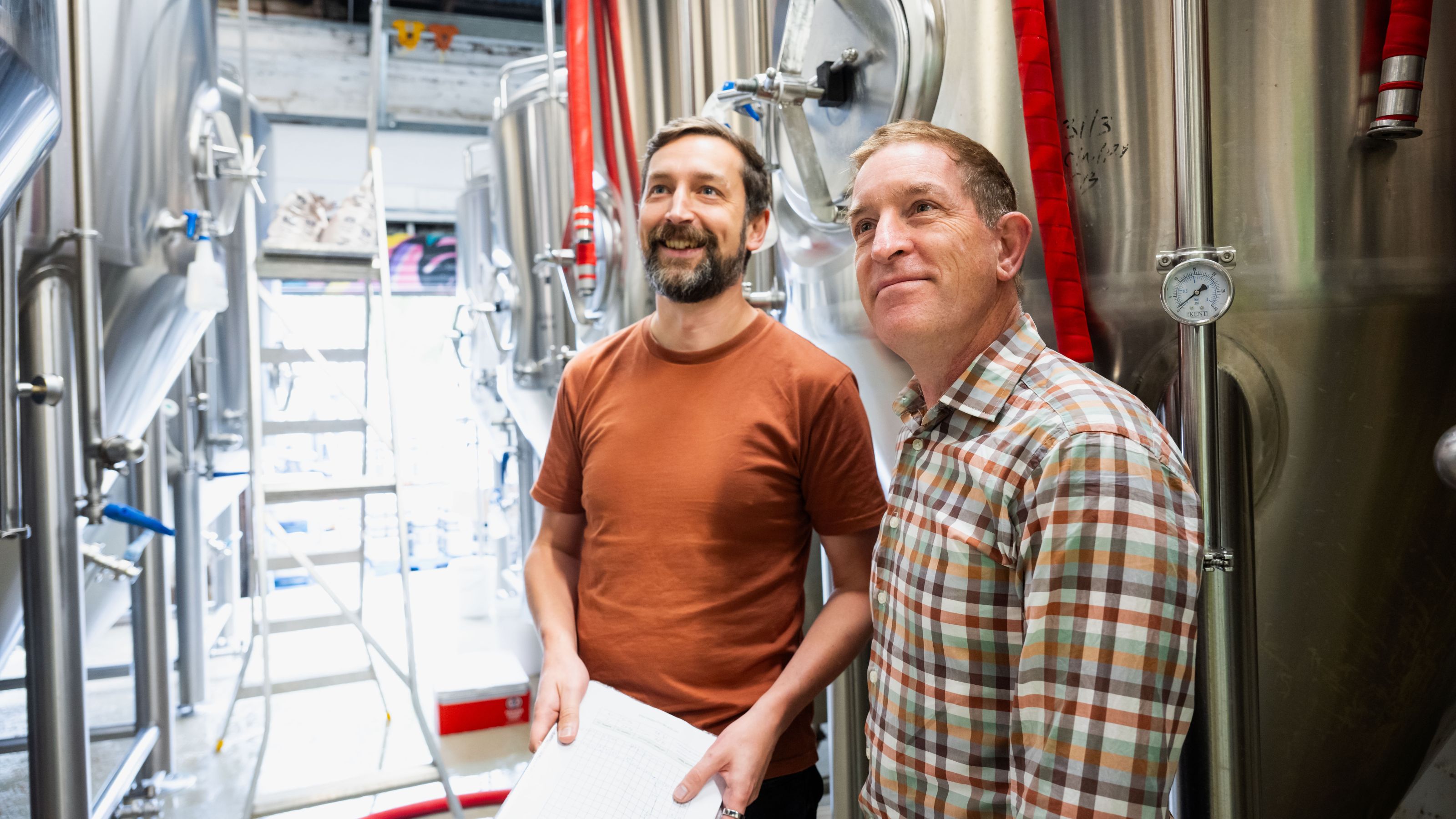 Peter Bircham and Simon Hinkley standing in front of a row of stainless steel brewing tanks inside a brewery.