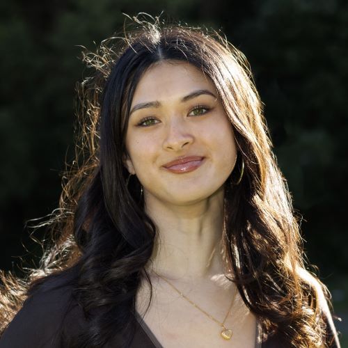 A person with long hair and a dark shirt, standing outside, smiling.