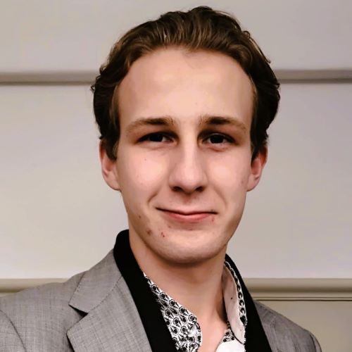 A smiling person standing in front of a beige background, wearing a grey jacket. 