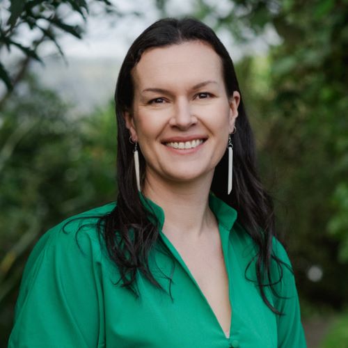 A person smiling and standing outside, wearing a green blouse and long white earrings, with trees in the background.