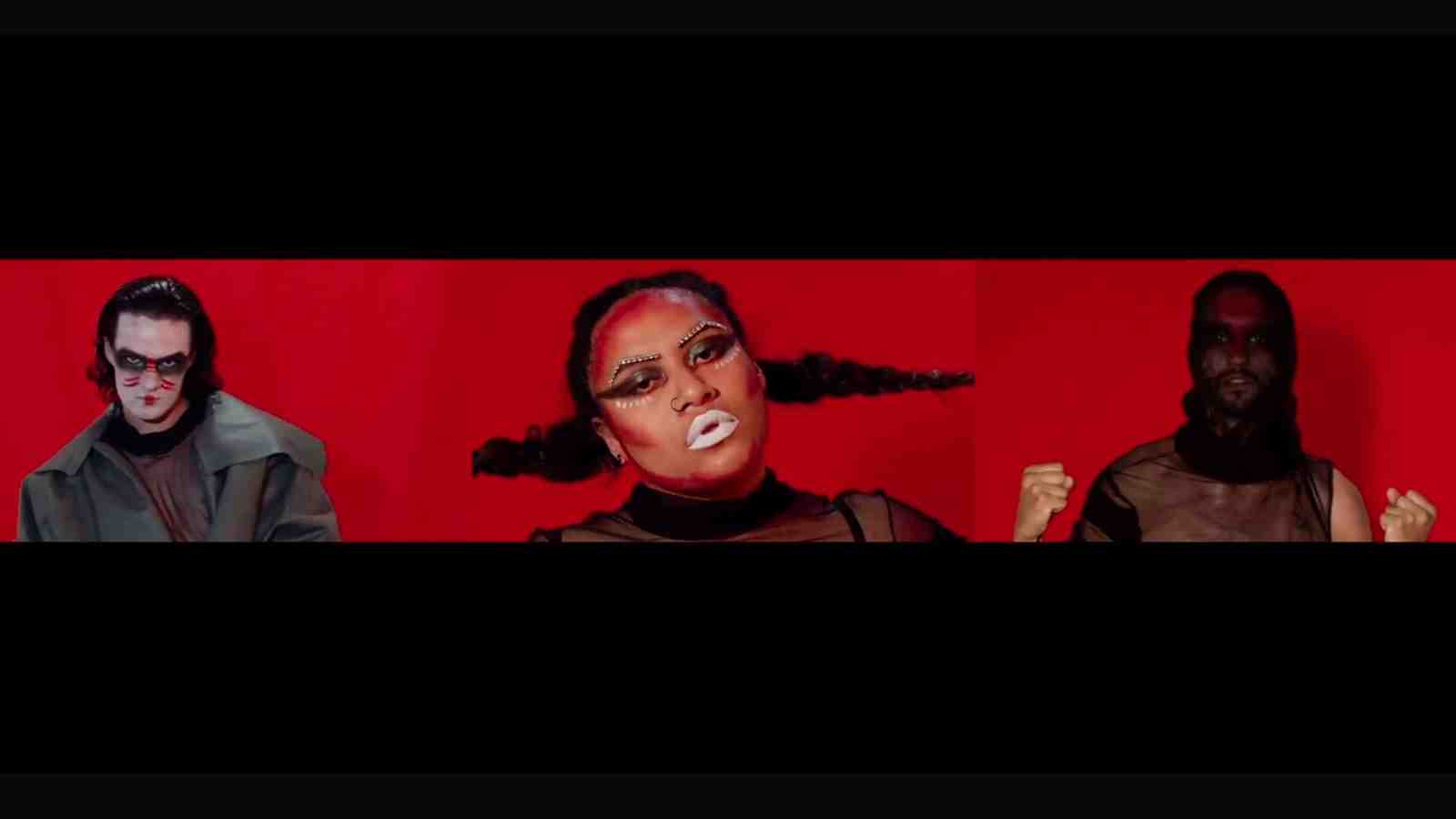 A still from 'Battle in Being' – Three performing artists in a red room wear elaborate makeup and dark clothing and hold intimidating facial expressions.