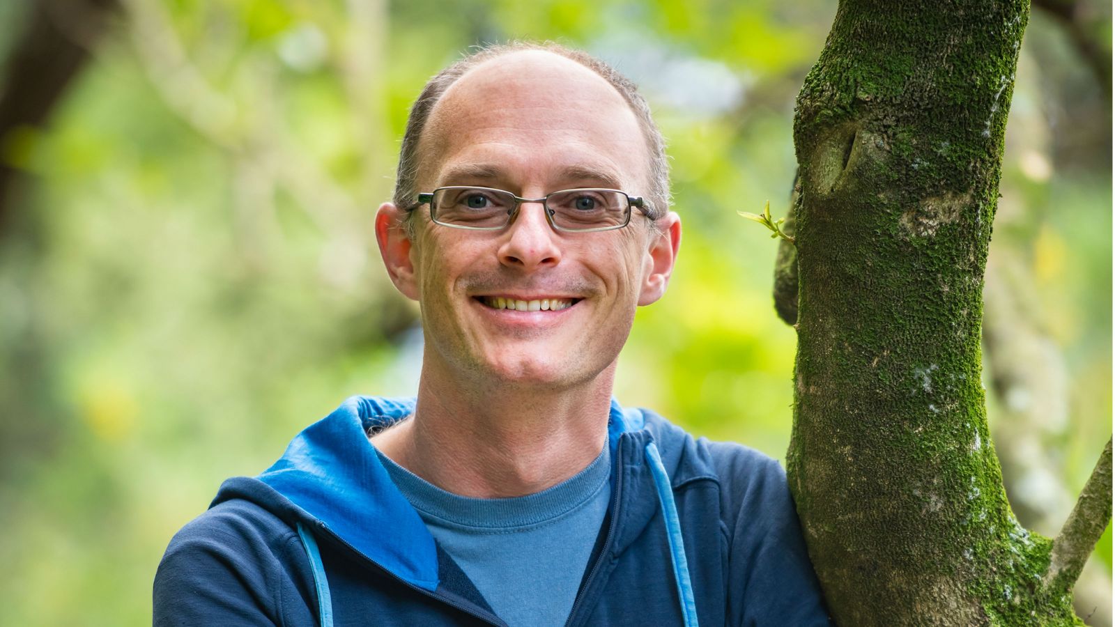 Stephen Marsland, Professor of Mathematics and Data Science, leaning against the mossy trunk of a tree. 