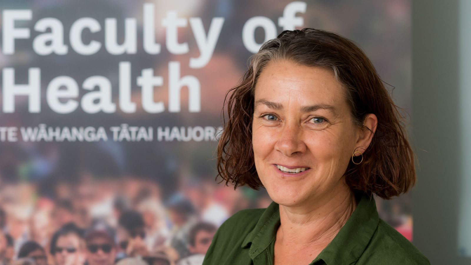 Dr Terry Fleming stands before a poster that includes the text 'Faculty of Health, Te Wāhanga Tātai Hauora' and a graphic of a crowd of people.