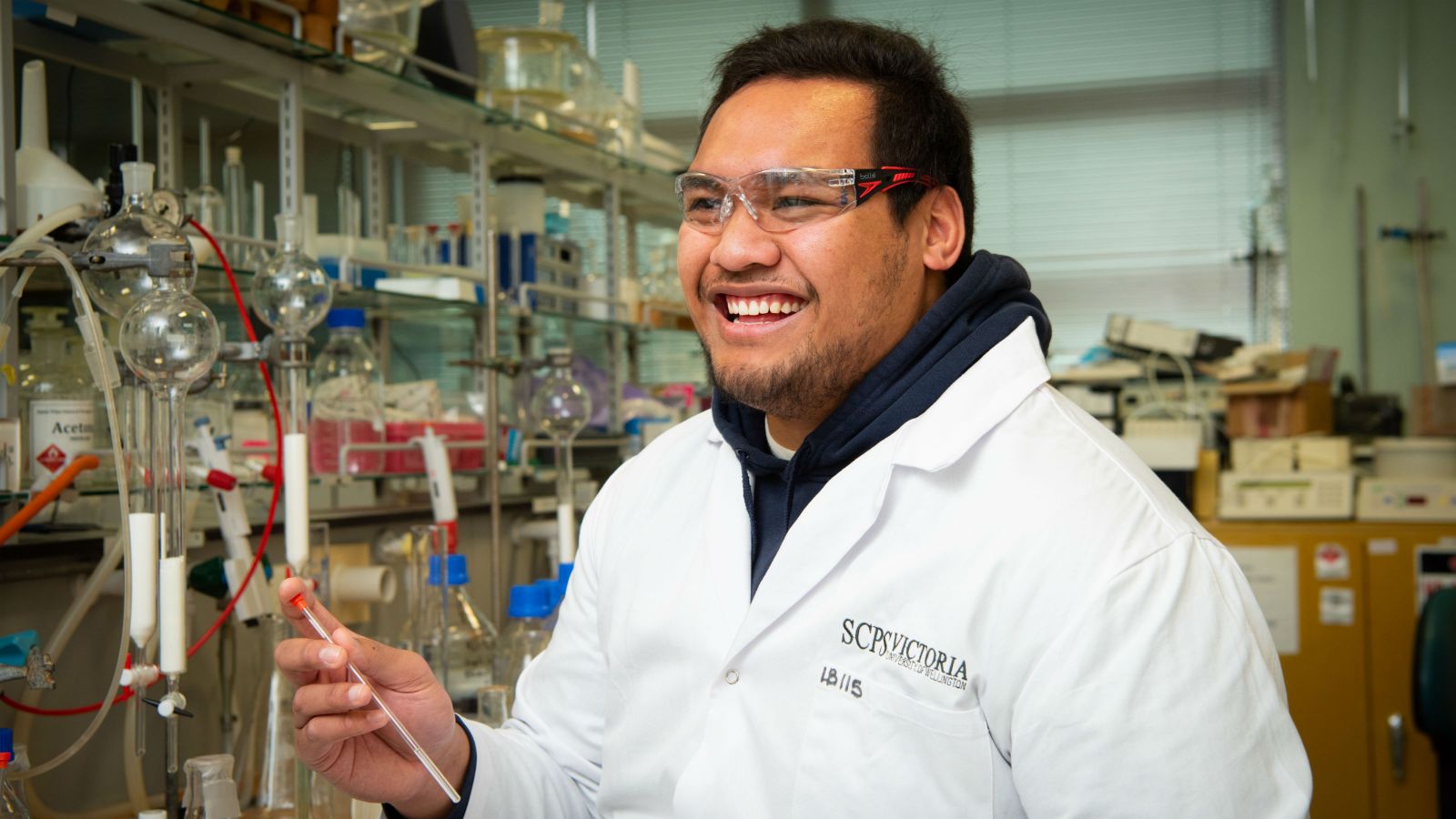 Brett, wearing a lab coat and safety glasses, laughs at something off camera. He is holding chemistry equipment with other equipment on the bench next to him. 