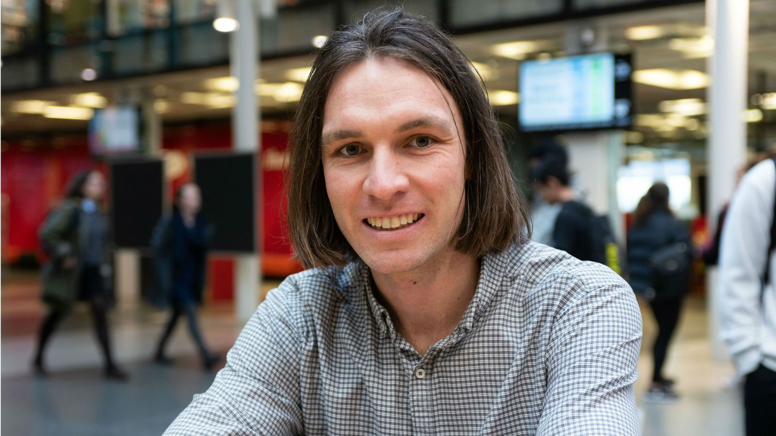 Ben sits at a table in the hub. Behind him is the blurred outline of people, digital screens and the library. 