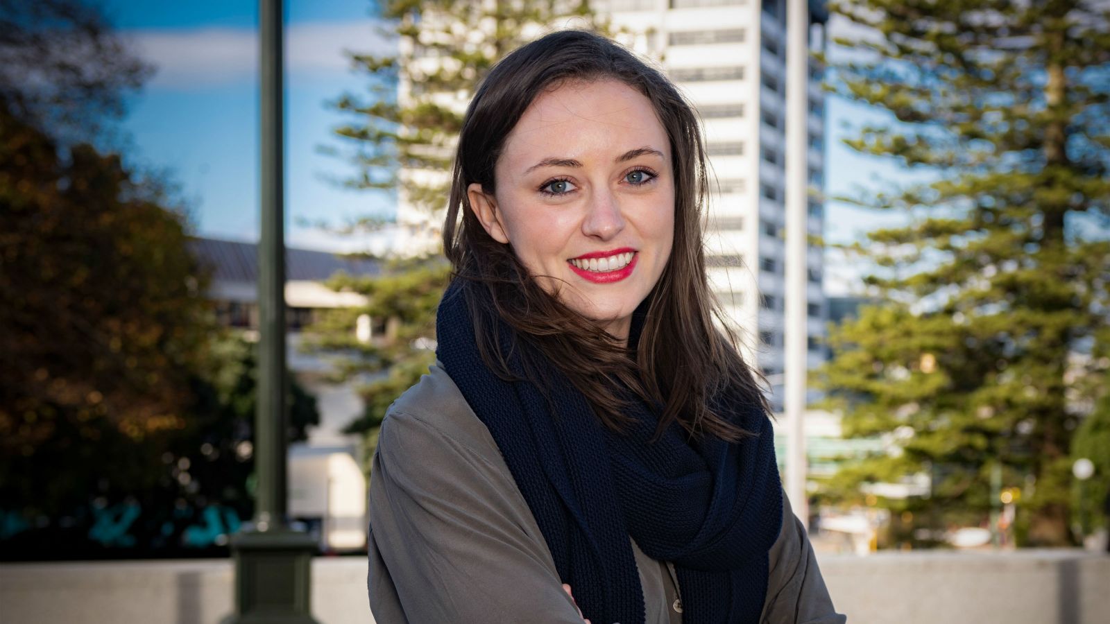 Kelle Howson, wearing a dark grey shirt and black scarf, stands with her arms crossed in Wellington city. Behind her is the blurred outline of a high rise building and trees.