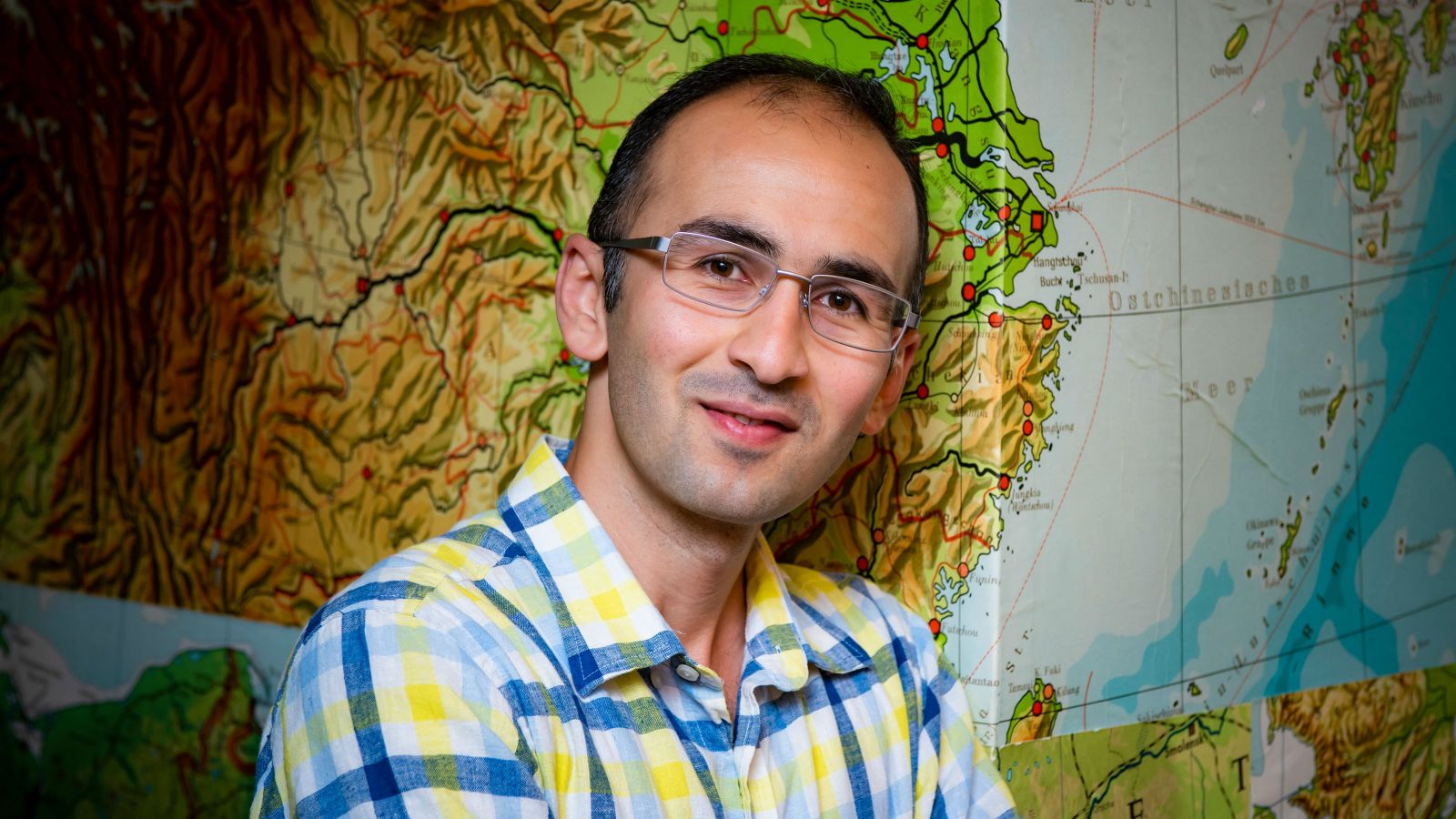 Omid leans against the wall and looks at the camera. The wall paper is a world map. 