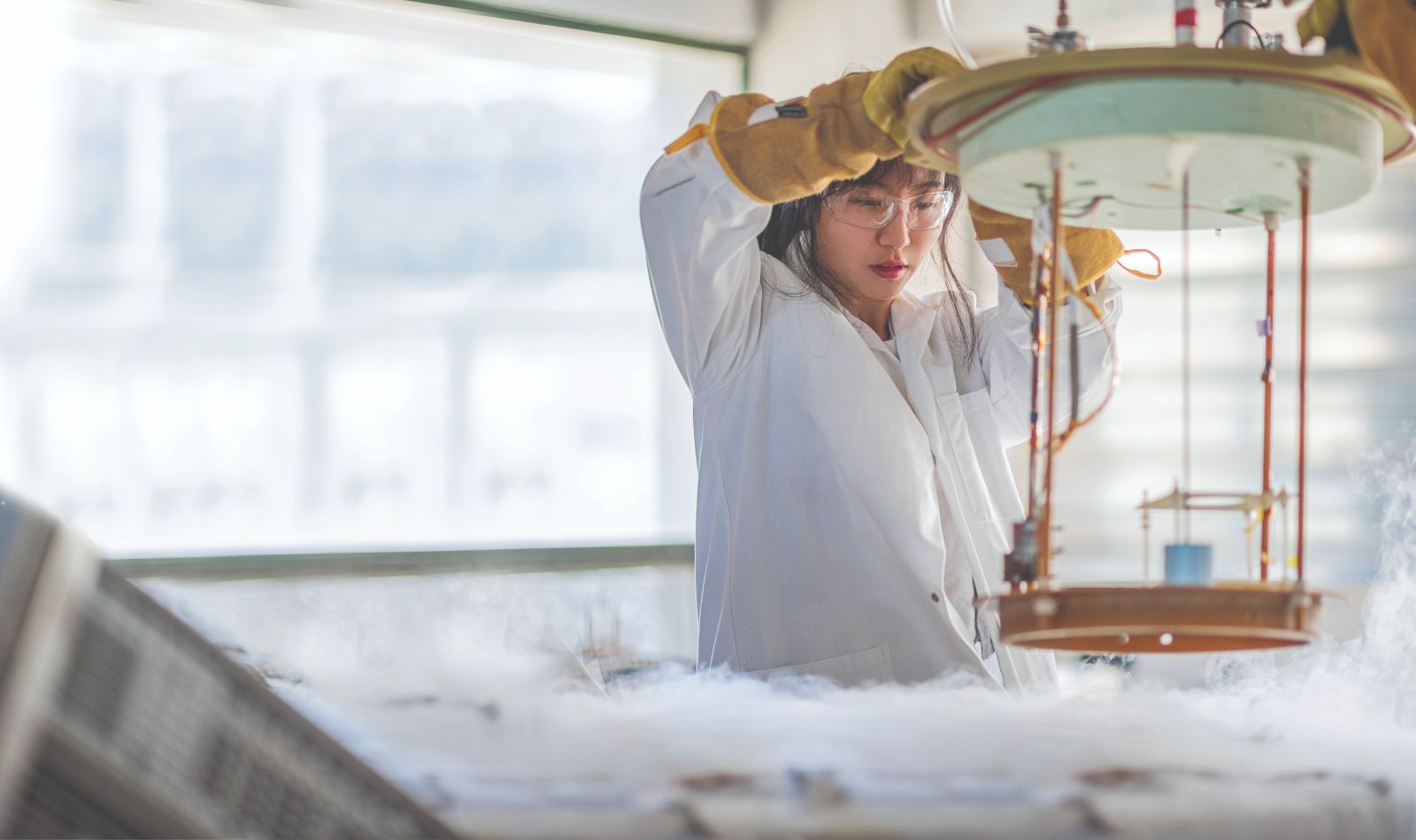 A female student, wearing safety glasses and a lab coat, holds up a beaker of blue liquid and looks at it. 