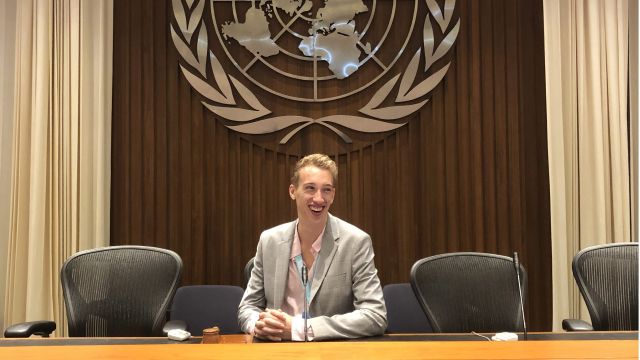 Luke White sits at a large wooden desk, the large silver United Nations logo on the wall behind him