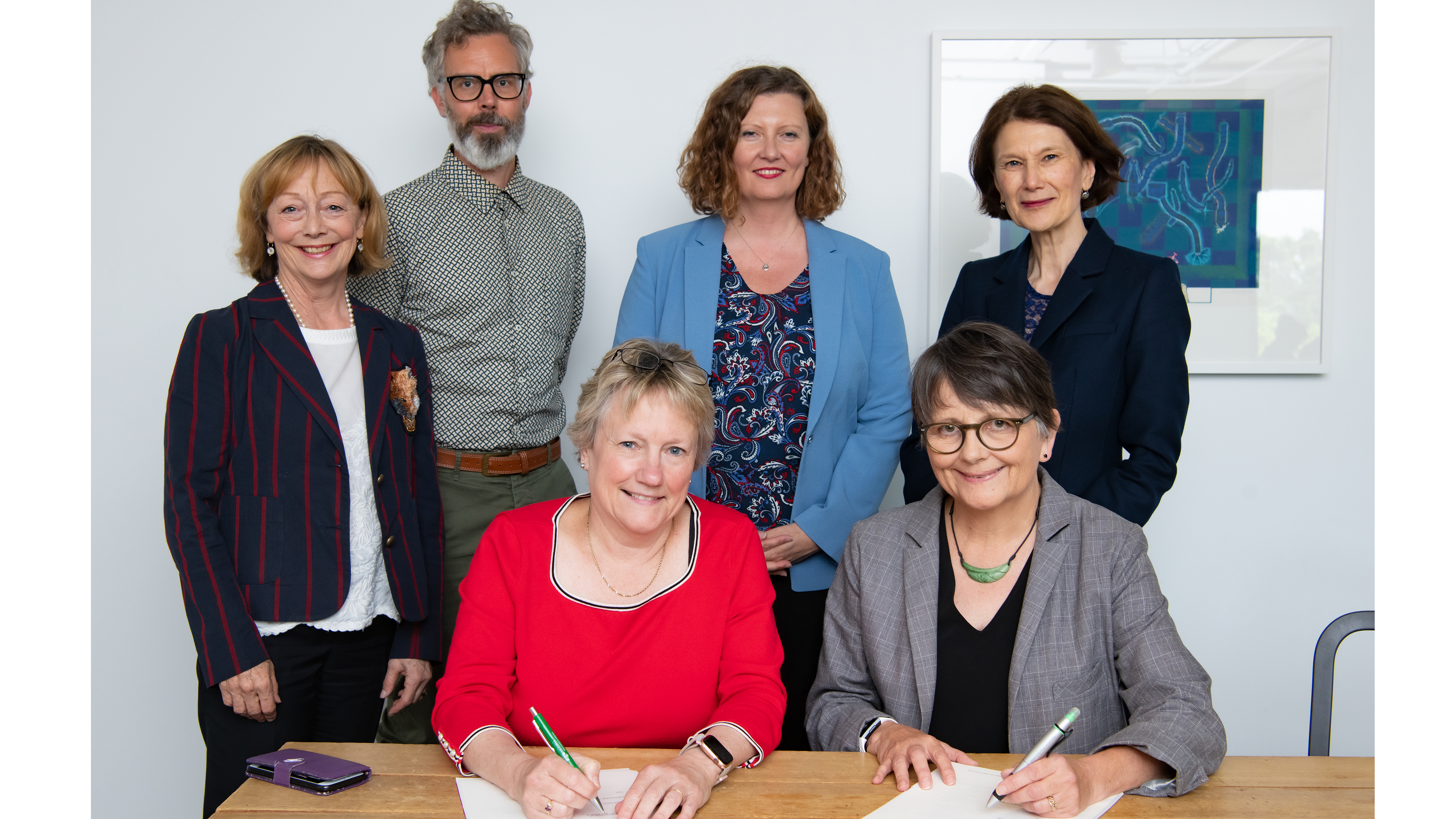 Two women sit at a table, each signing a document posing for the camera. Four other peoples, 3 women and 1 man, stand around them. Standing (L-R): Rosalene Fogel (Development Office), Dr Dugal McKinnon (NZSM), Steff Kemp (CMNZ), Professor Jennifer Windsor (PVC, FHSS and FoE). Sitting (L-R): Patricia Danver (ED, Victoria Foundation, Development Office), Catherine Gibson (CE, CMNZ).