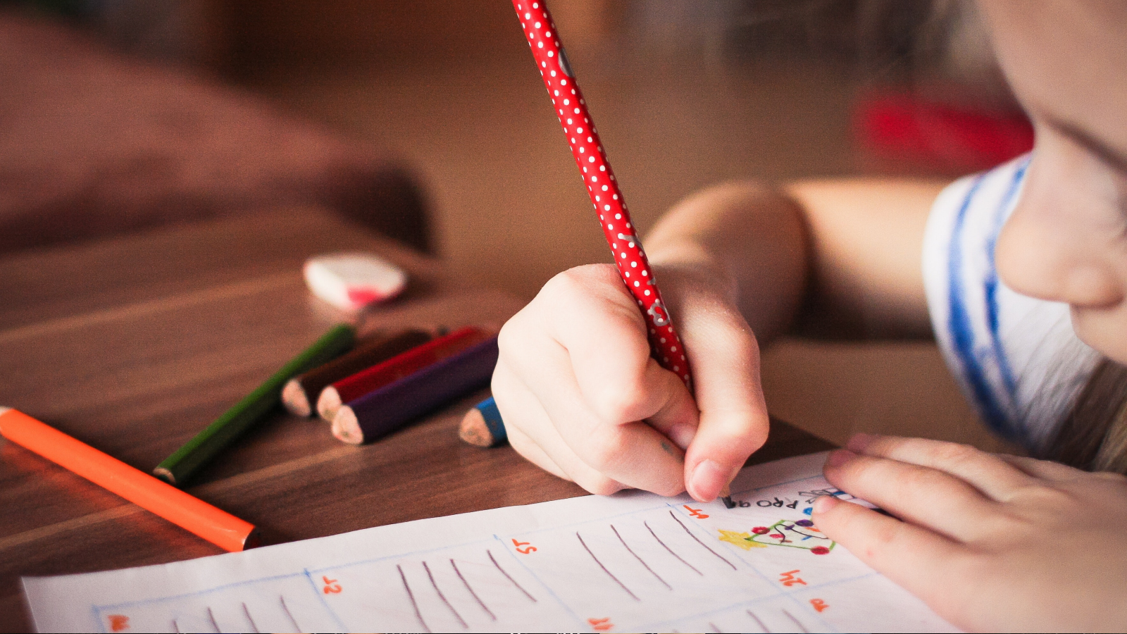 A child writes with a red pencil at a table.