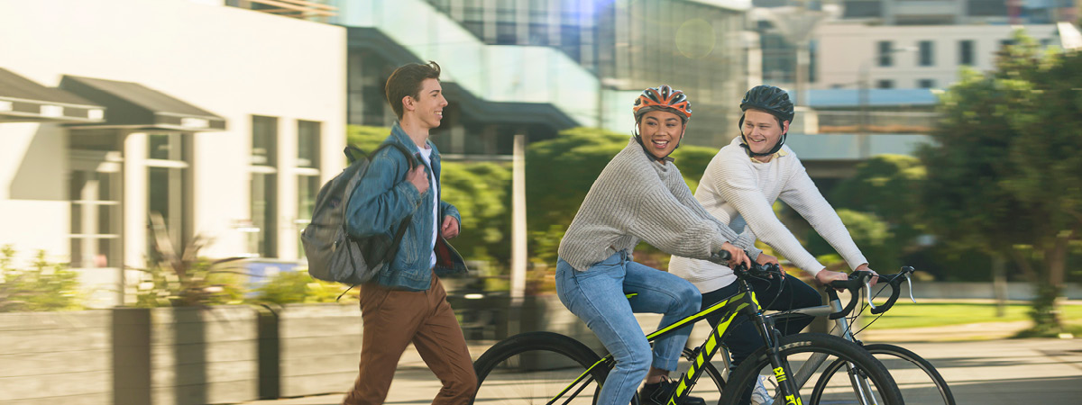 Get in touch banner – Two students on bicycles smile while passing another student who is walking. banner image