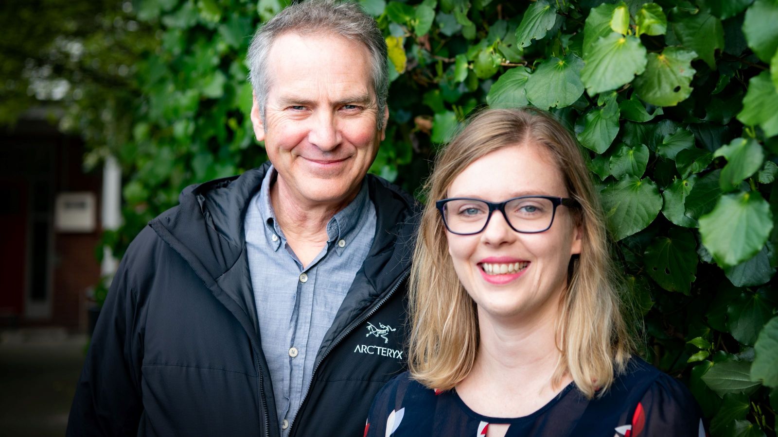 Jeff and Hannah stand in front of a large green bush.