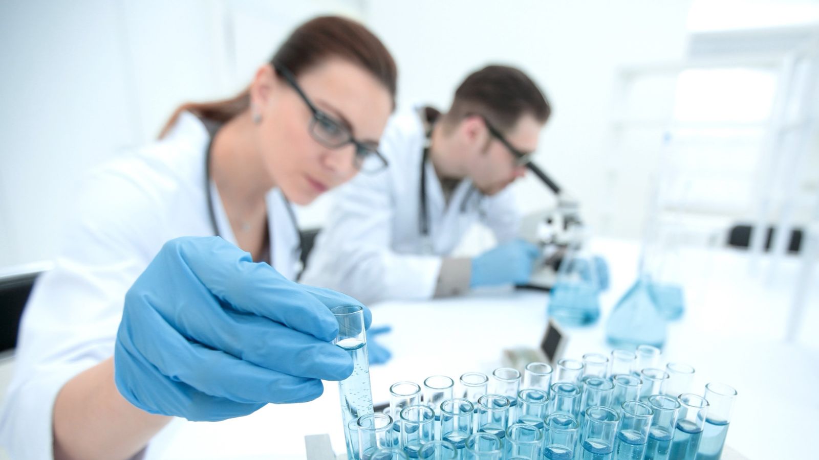 Two white-coated researchers in a laboratory using a microscope to look at test tube samples
