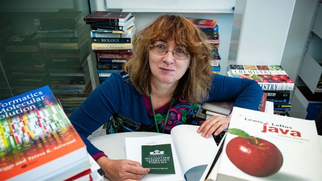 Sue Hall sits at a white table surrounded by stacks of books. She holds an open book and a Victoria University of Wellington card between her hands. 
