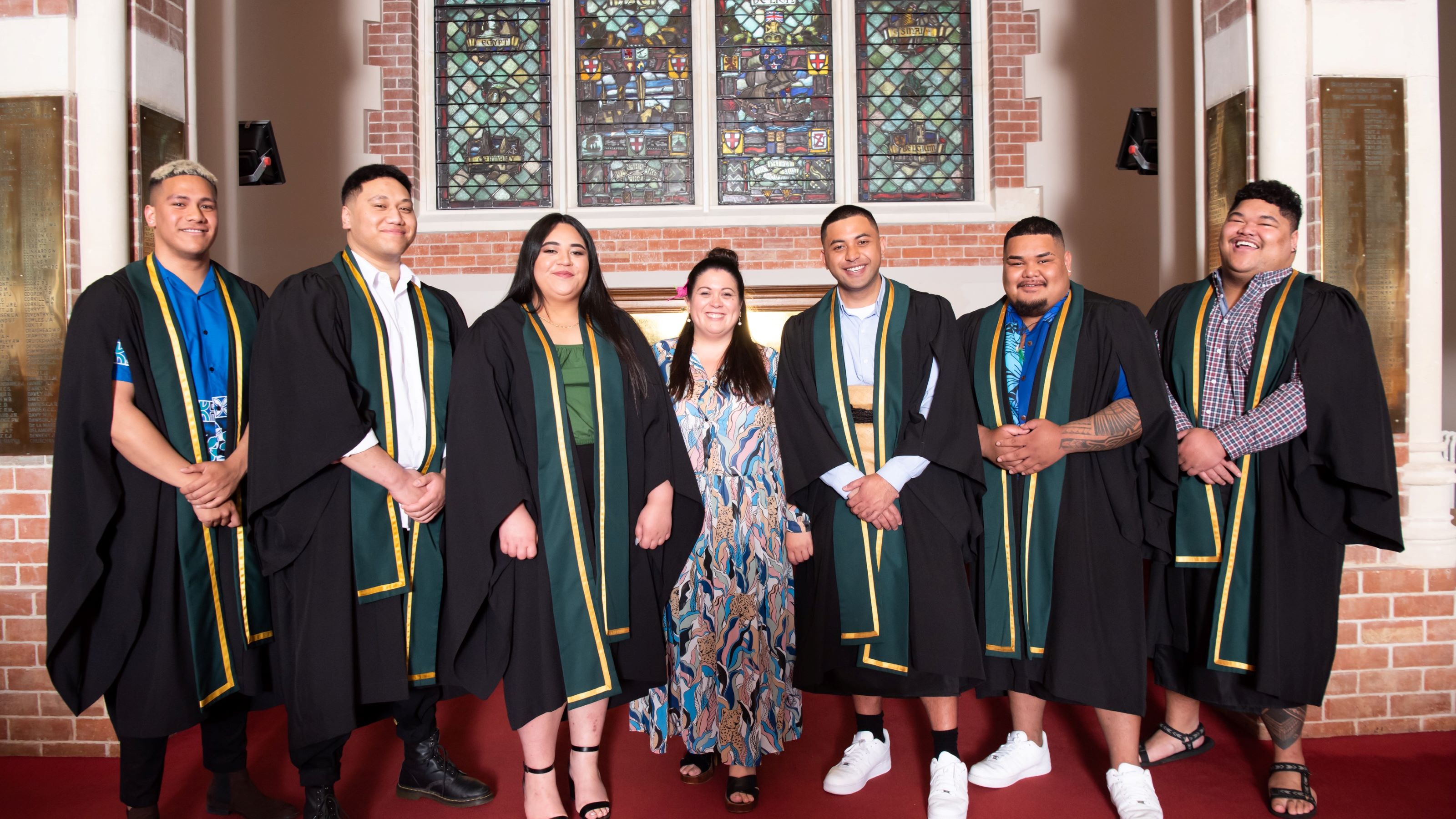 Pasifika students in academic dress (robe and sash) stand in front of a red brick wall with stained glass windows