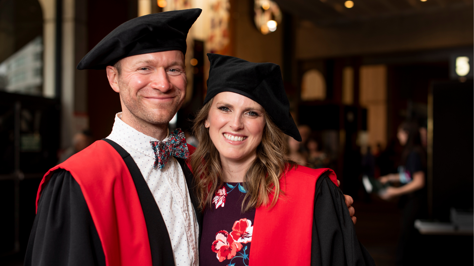 Male with bowtie in phD red robes, female with floral dress in PhD red robes