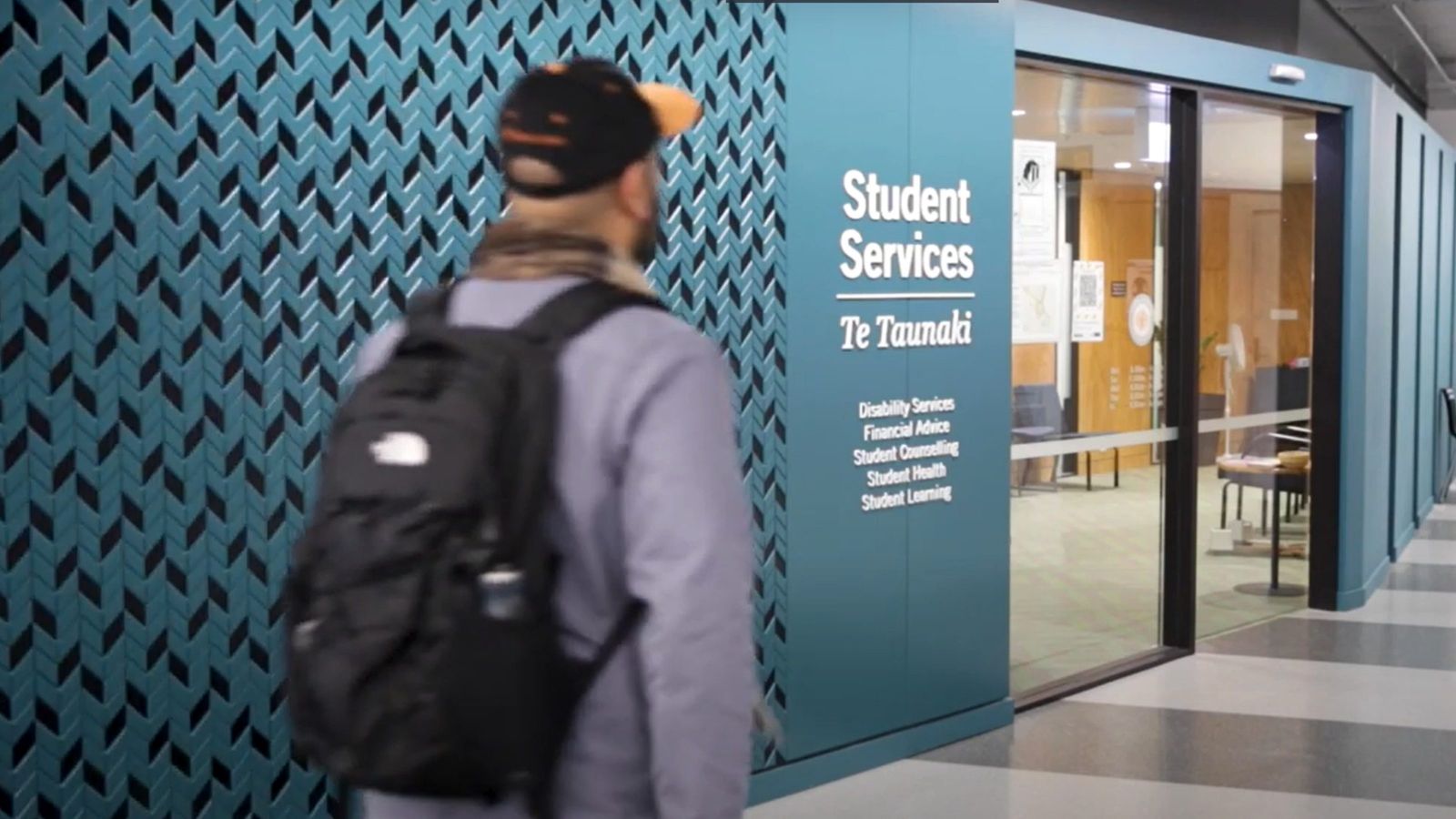 Frame from a video showing a young man wearing a backpack walking towards the Student Services office at the University.