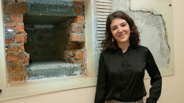 Rachel stands beside an exposed red brick wall which has a deep hole in it.