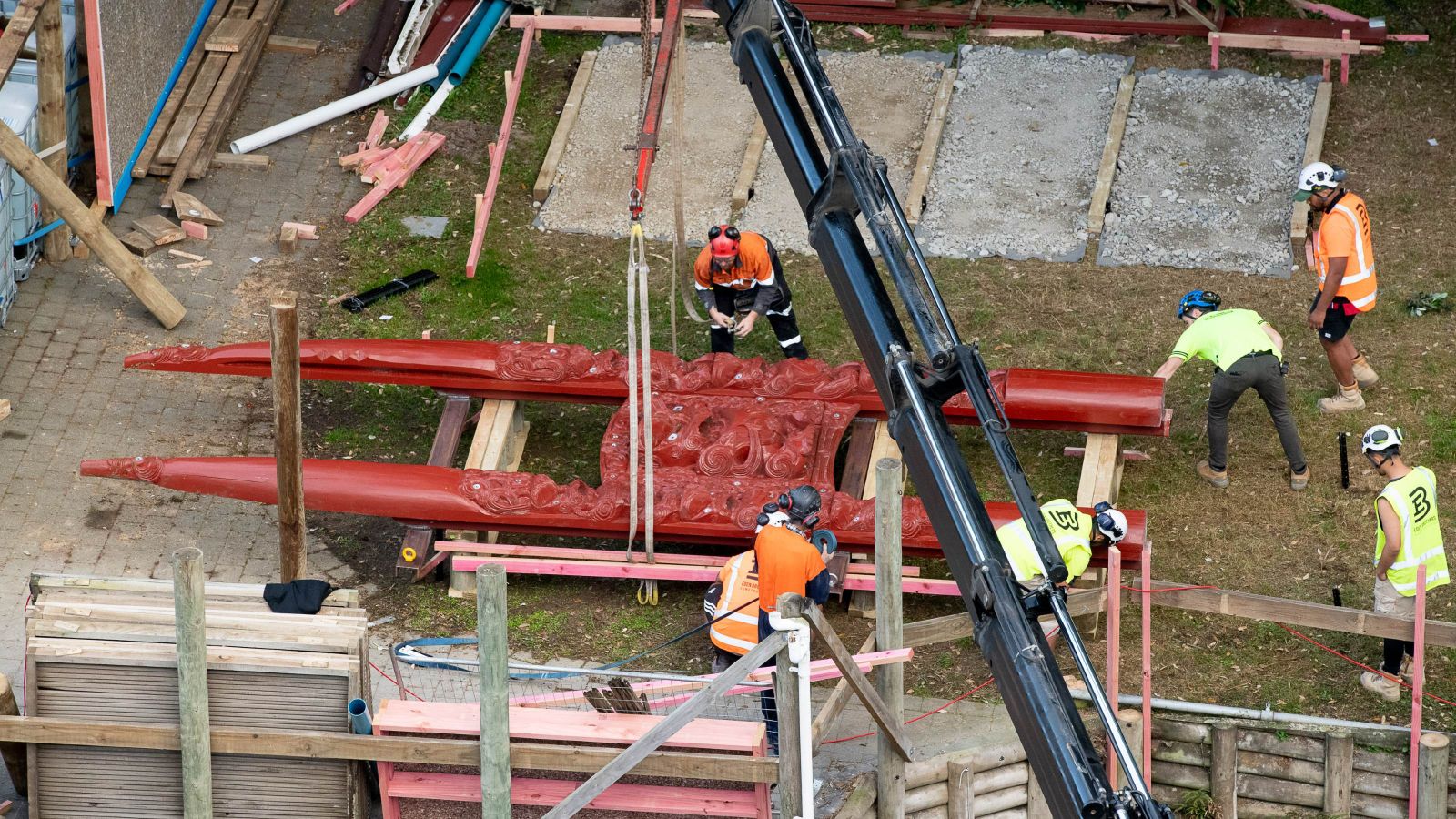 An aerial view of a carved waharoa (entranceway) being winched by a crew of construction workers in high vis vests and laid on the ground.