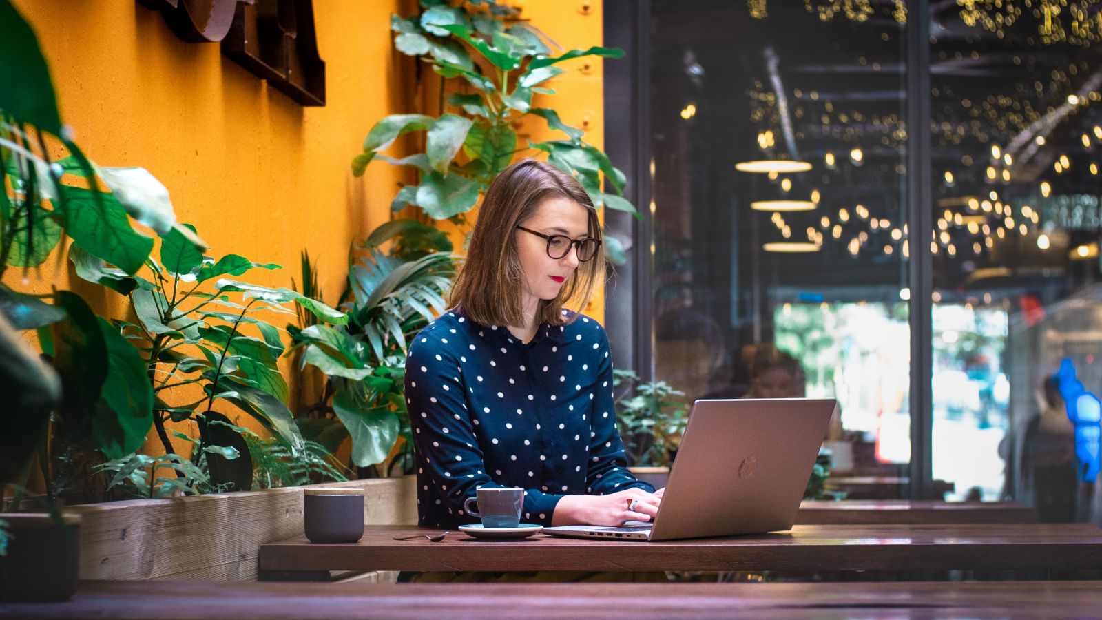 A student with glasses sits at a coffeeshop looking at her laptop.