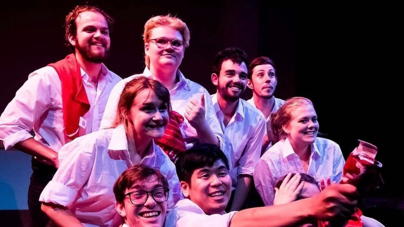 Eight actors on stage, wearing white shirts reflecting pink light, gathered together, facong the public and smiling.