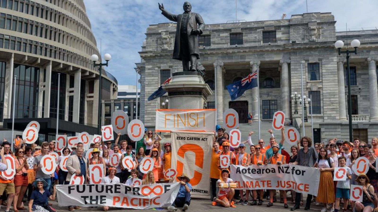 Large group of students holding orange '0' signs and banners saying 'Future proof the Pacific' and 'Aotearoa NZ for climate justice', with the Beehive and the statue of Richard Seddon in the background.