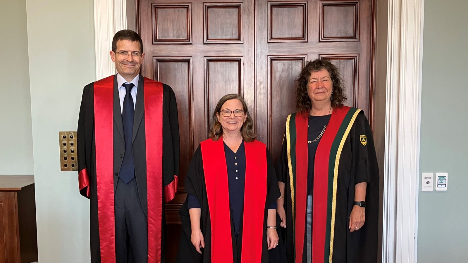 Three people in academic garb pose in front of a polished wood panel wall.