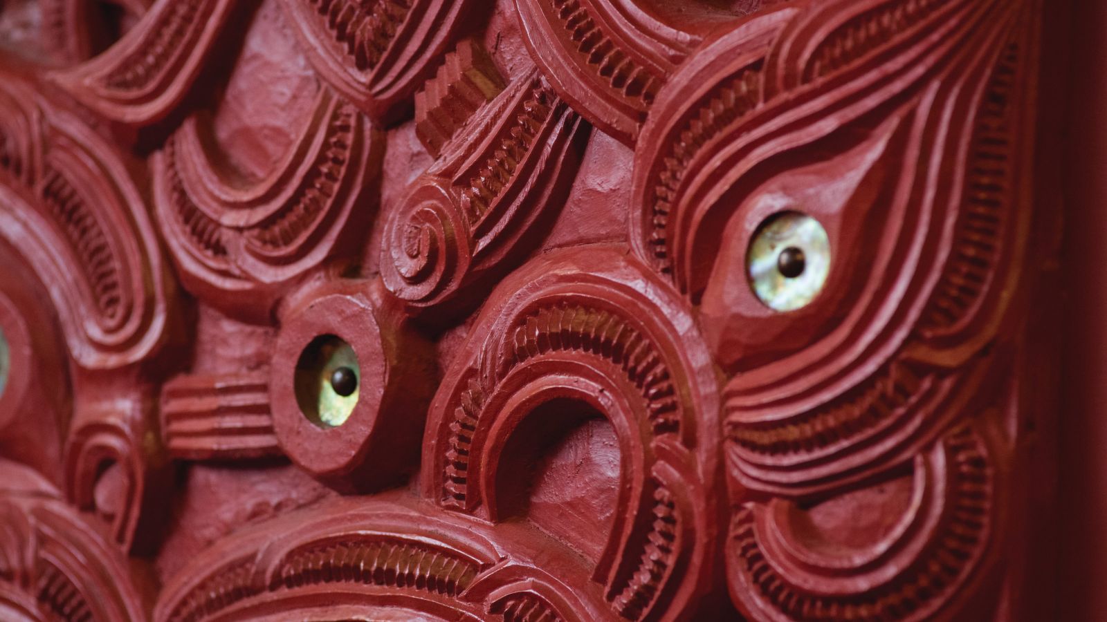 Red painted carved panel detail, with curved lines and two paua shells inset as eyes.