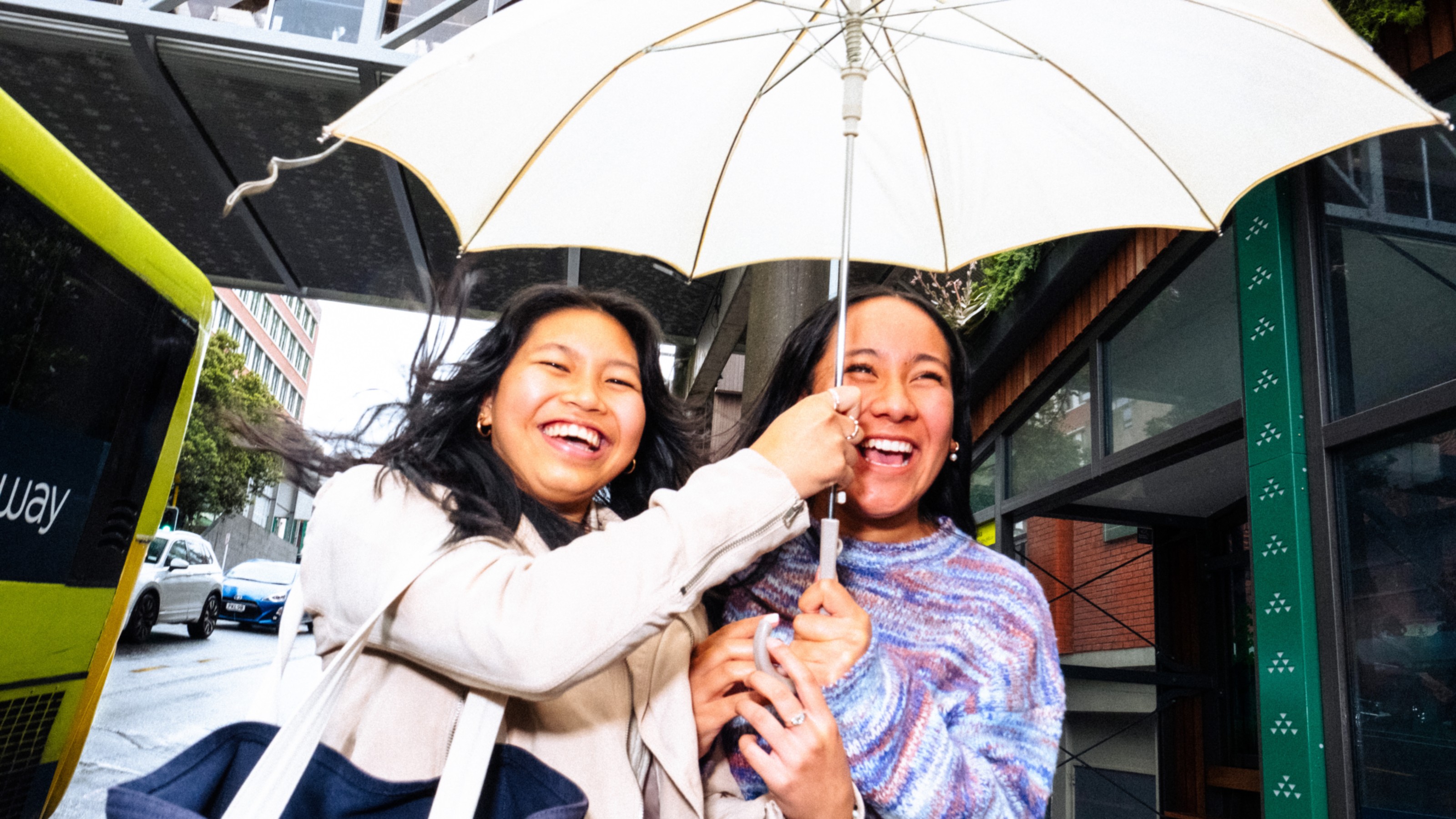 Two people under a white umbrella on a rainy day, near a green bus and urban buildings; one wears a light coat with a dark bag, the other a colourful jumper.