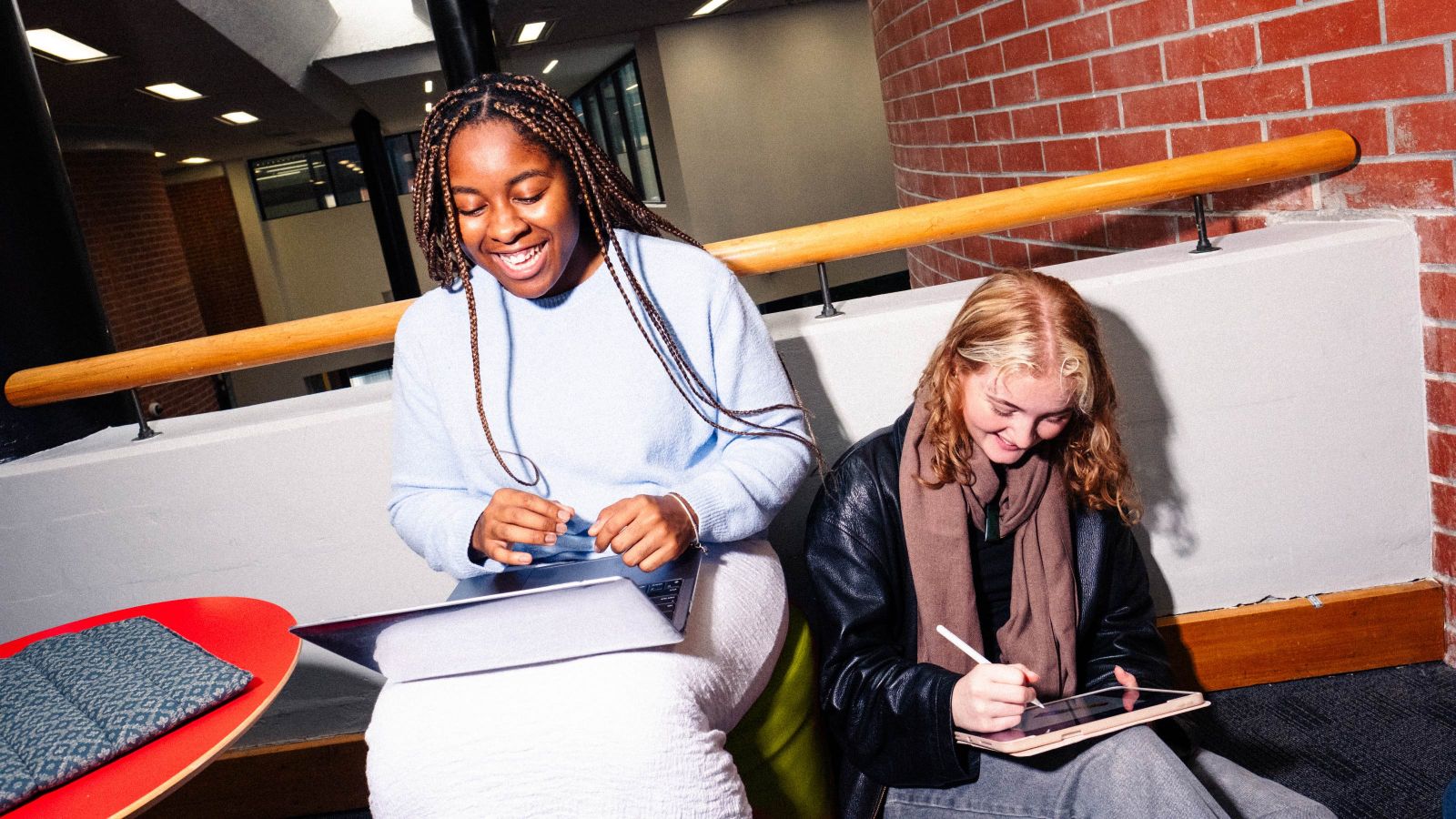 Two people sit indoors; one holds a laptop, the other uses a tablet with a stylus.