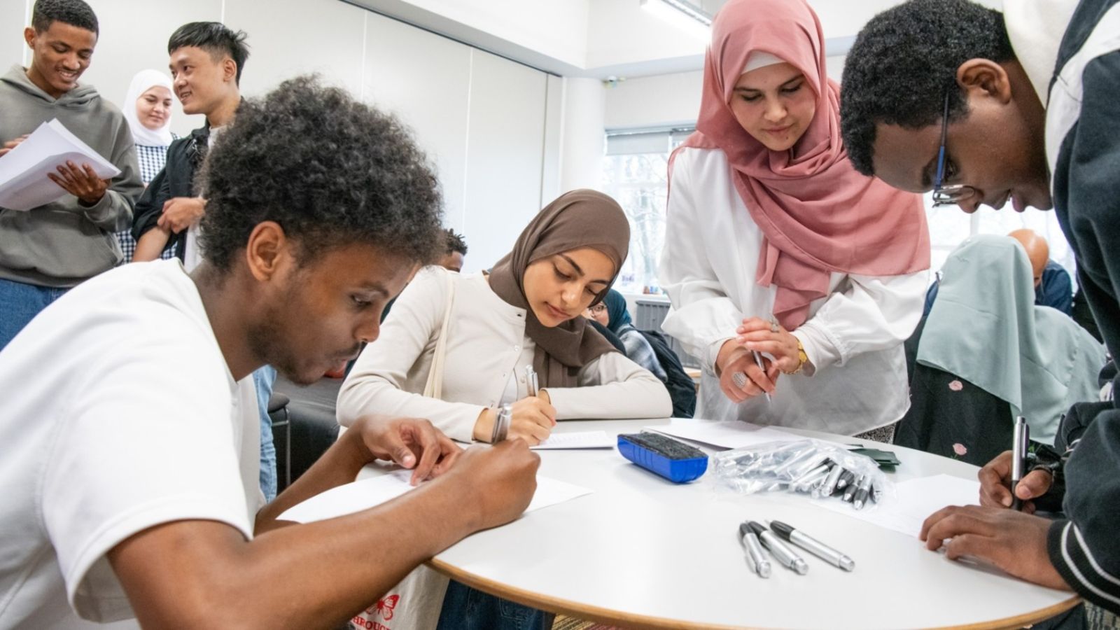 Two male and two female students are at a table studying. The female students are wearing hijabs. All students have pens in their hands and are focused on the paper they are writing on. Two students are sitting on the left side and the other two students are standing on the right side.