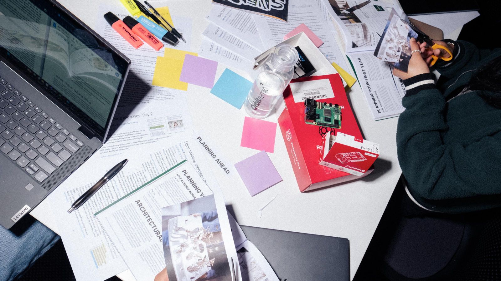 Aerial view of a table with an open laptop, strewn with paper and we can see students cutting paper with scissors 