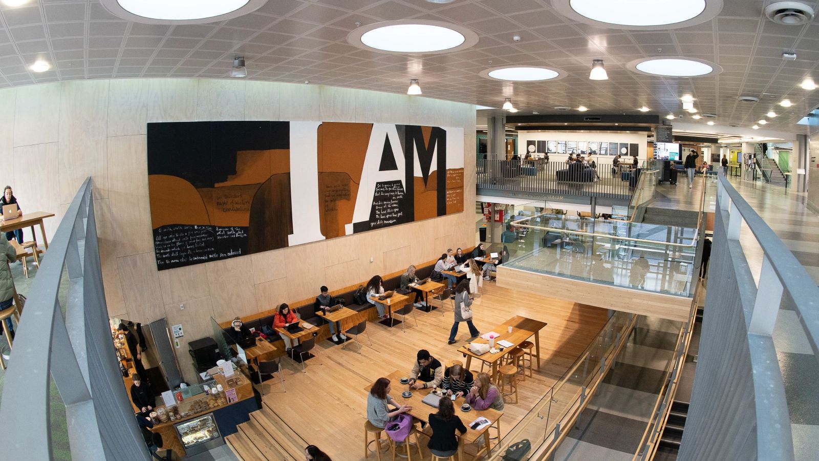 Students sitting and studying in a bright atrium with a large ‘I AM’ mural on the wall.