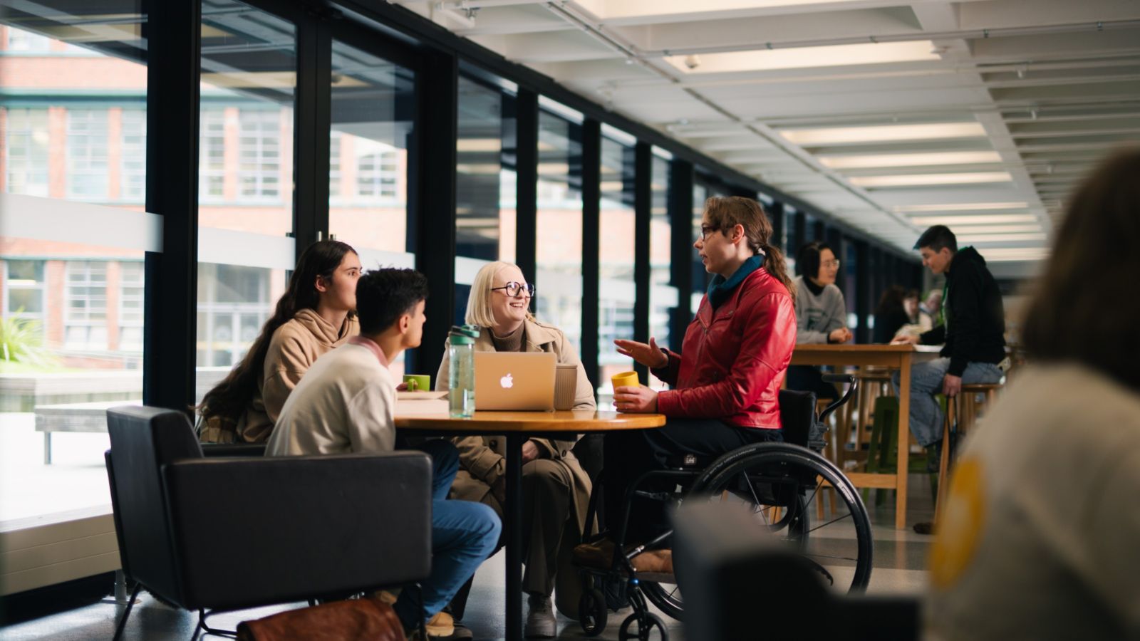 4 students sitting at a round table with laptops and cups of coffee. One student is in a wheelchair