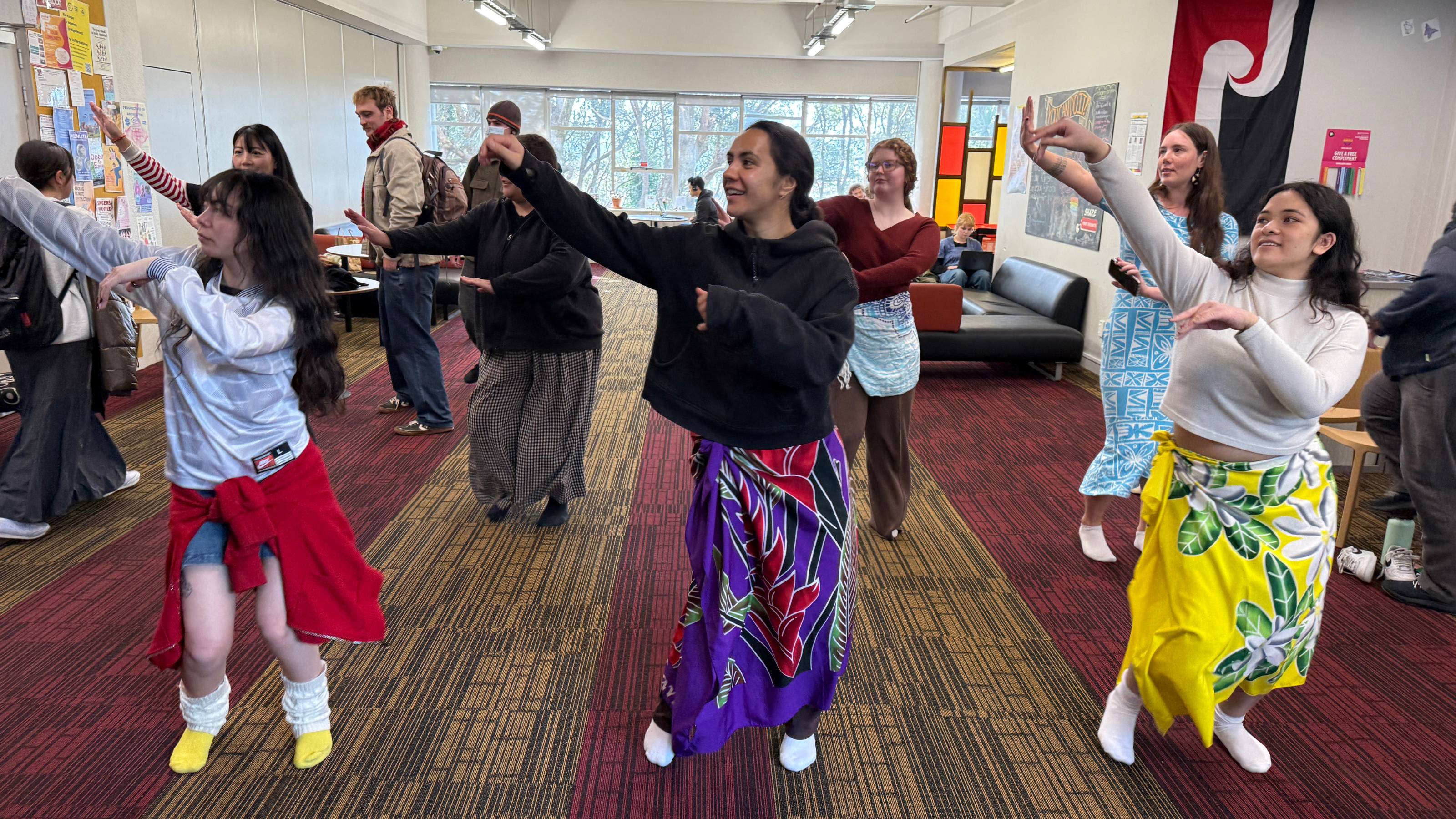 A group of students standing in rows indoors, following the same dance movements with raised arms. Several students wear colorful patterned skirts tied around their waists, while others wear casual clothing. Chairs, couches, posters, and windows are visible in the background of the campus common area.