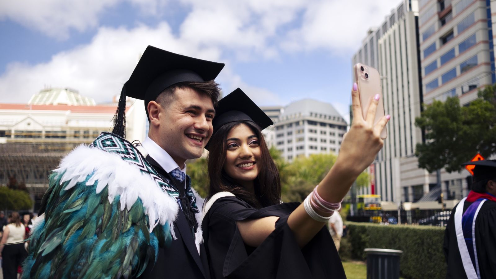 Two graduates in caps and gowns take a selfie outdoors during a graduation event.