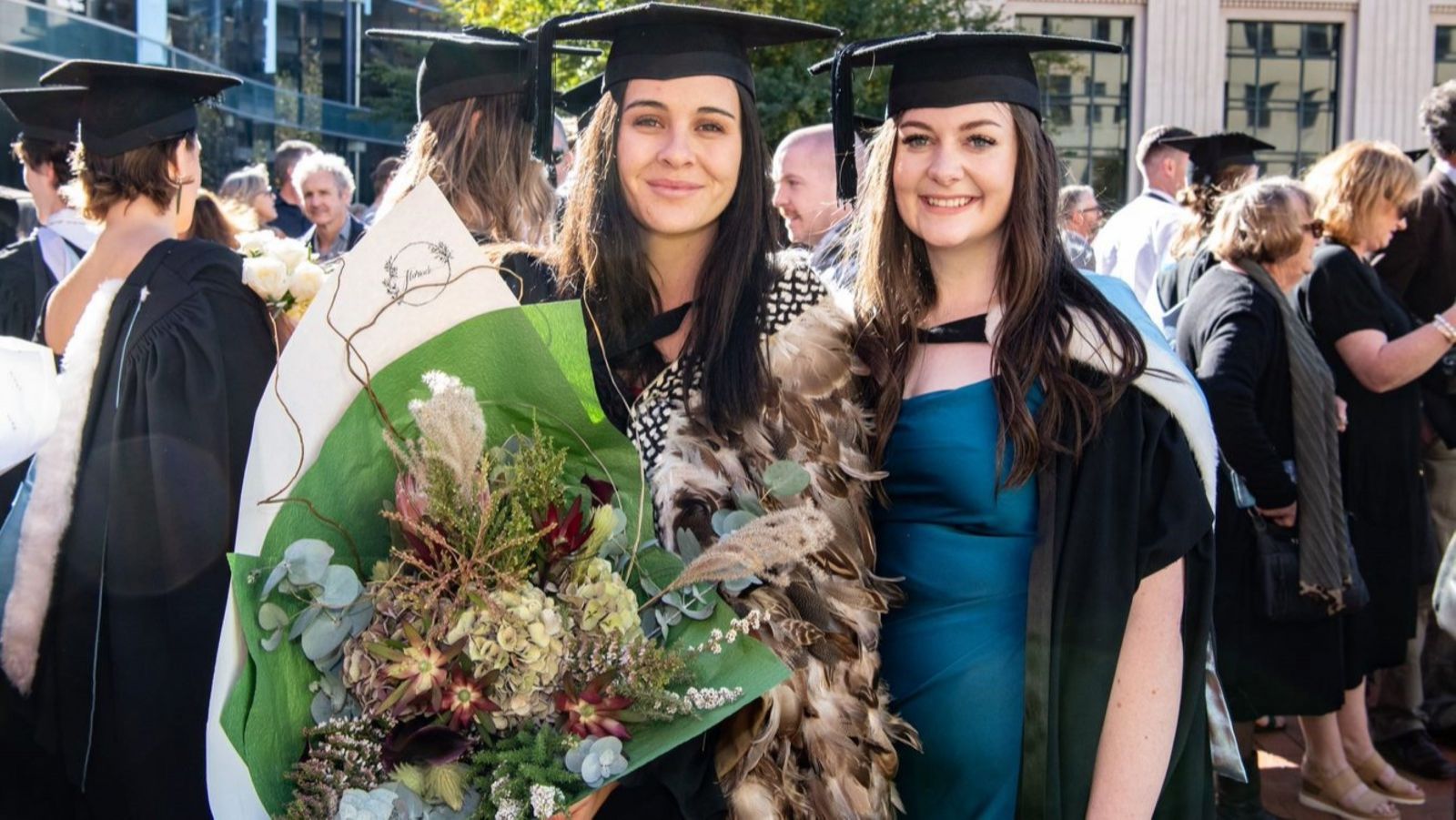 Two young women graduates, one wearing a feather cloak and holding flowers, in front of a crowd of other graduates.