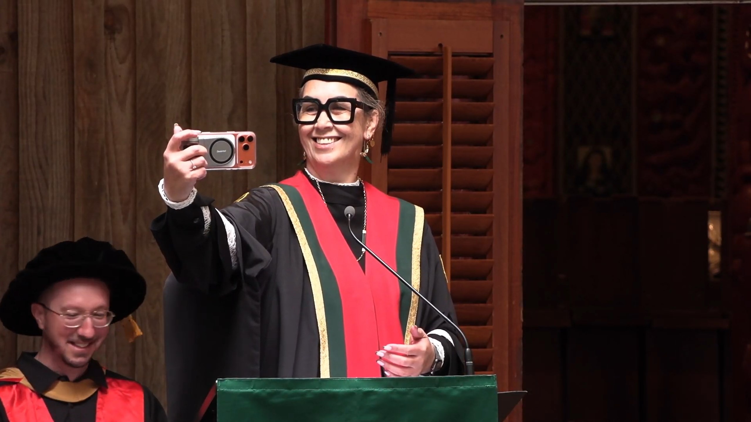 Person in graduation regalia holding a phone and taking a selfie at a podium.