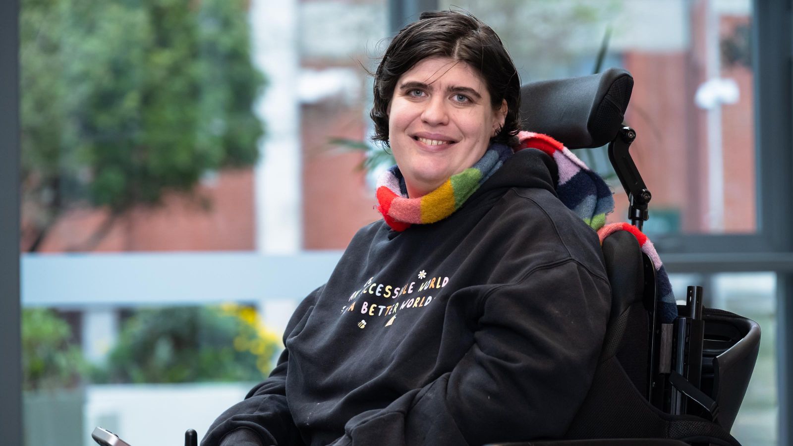 Rainbow Support Navigator, Etta Bollinger, smiling and wearing a rainbow scarf and a sweatshirt that says: An accessible world is a better world.