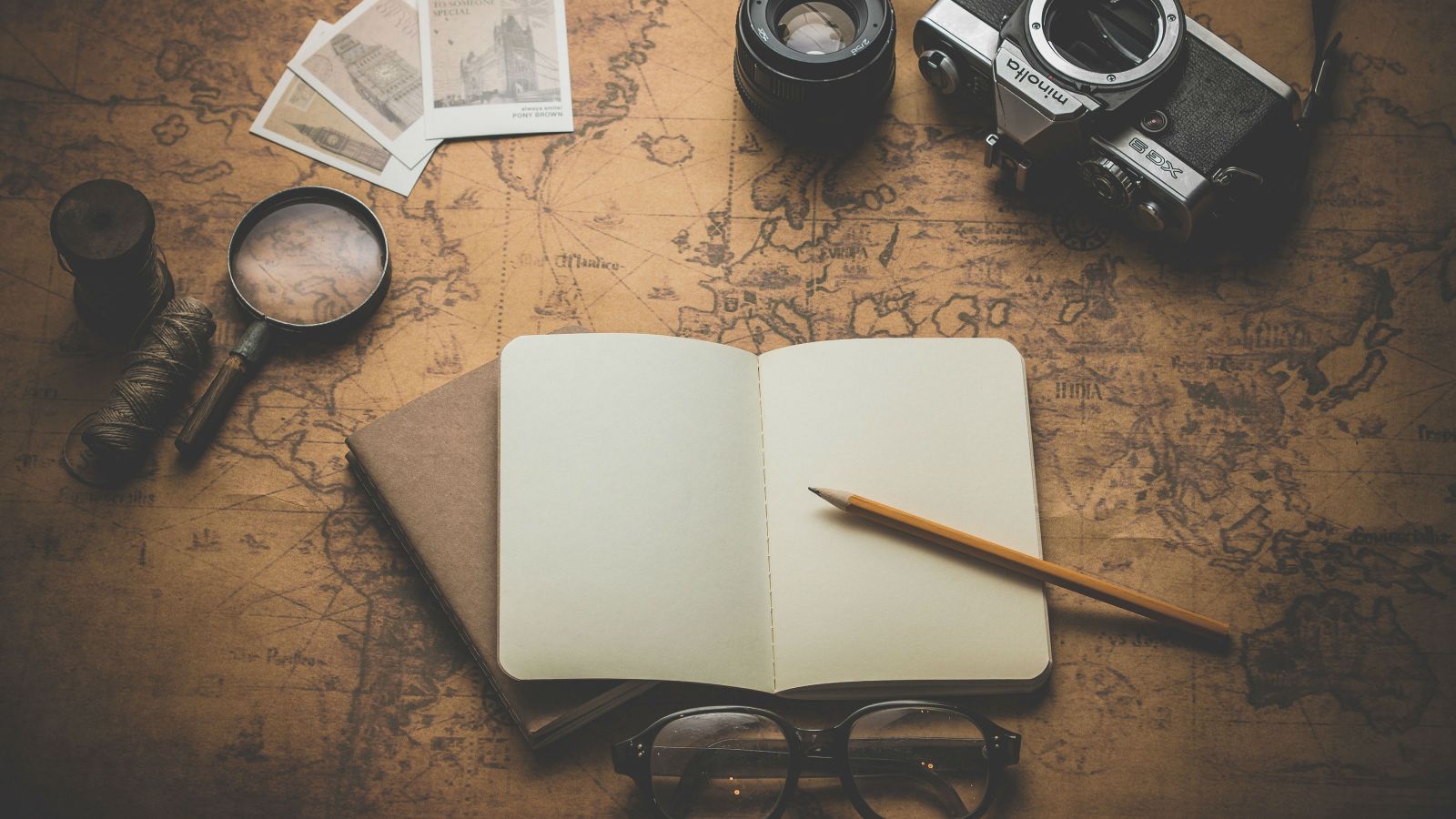 A notebook, pencil, magnifying glass, spectacles, camera and polaroids on a background of an old school world map.