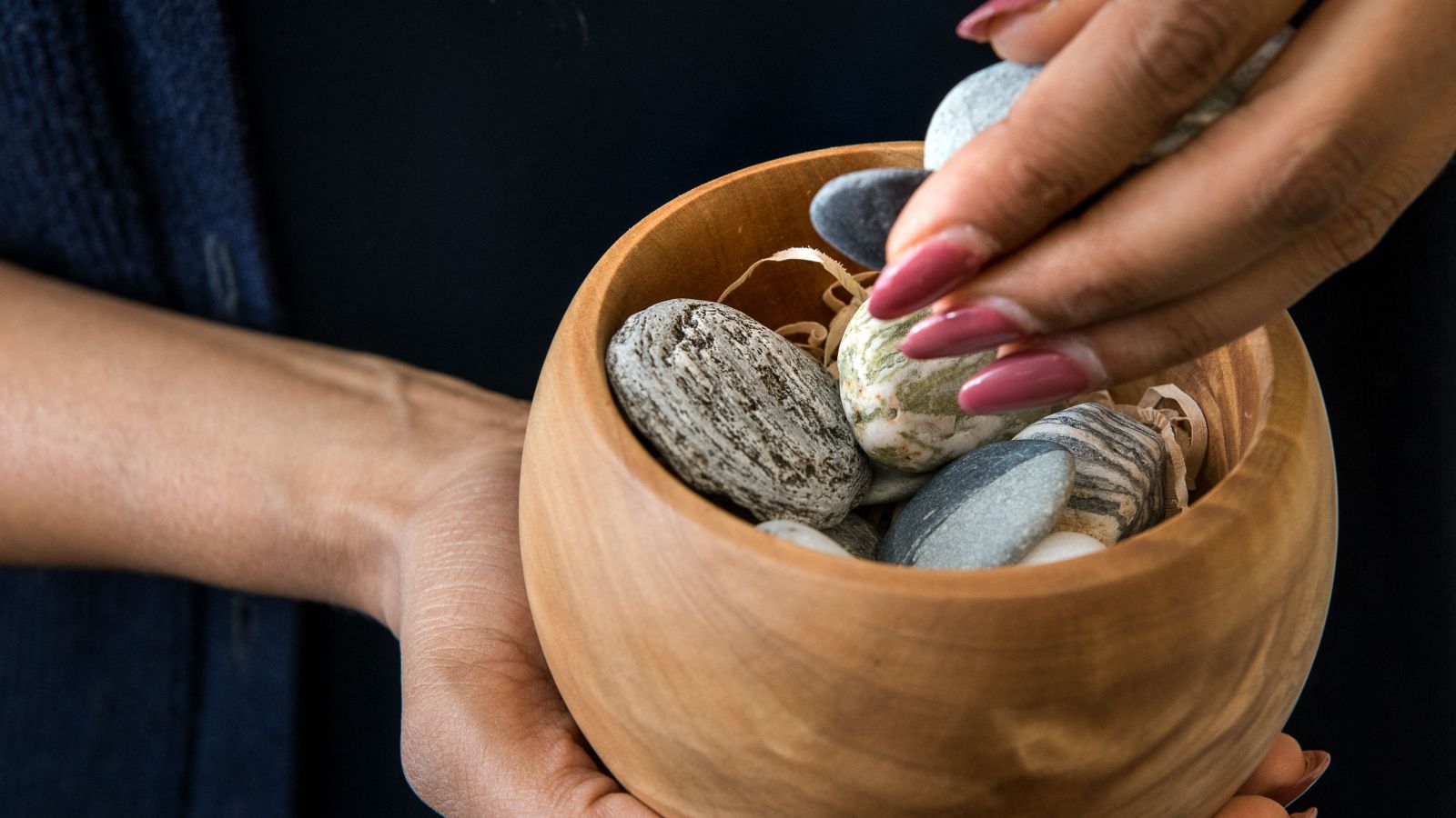 A woman holds a wooden bowl containing stones while she extends one of the stones as a gift.