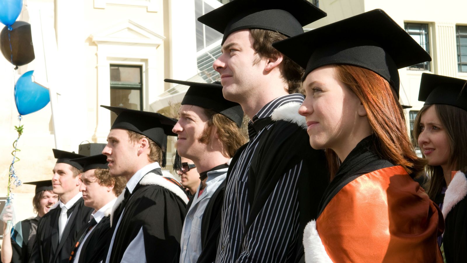 Graduating students stand in full wardrobe listening intently, a student at the end joyfully holds onto celebratory balloons.