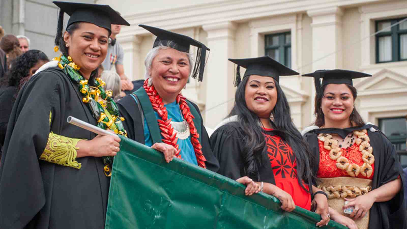 Three students and a professor all smile while wearing graduation gowns and caps, all holding a banner for a parade.