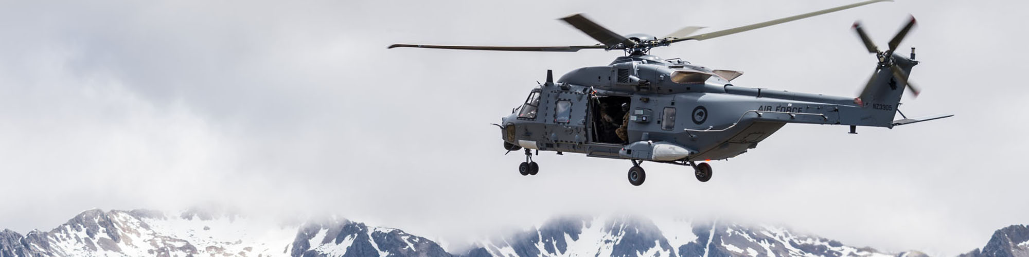 A large military helicpoter hovers in front of mountains under a cloudy sky